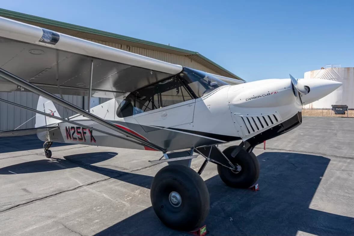 A close-up three-quarter front view of the white and grey Carbon Cub FX-3 aircraft N25FX showing the engine cowling and large tires.