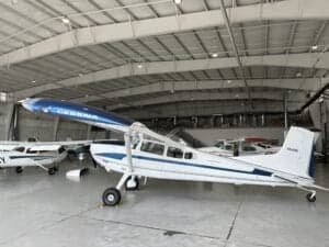 A Cessna aircraft with a white and blue paint scheme is parked in a hangar with other aircraft.