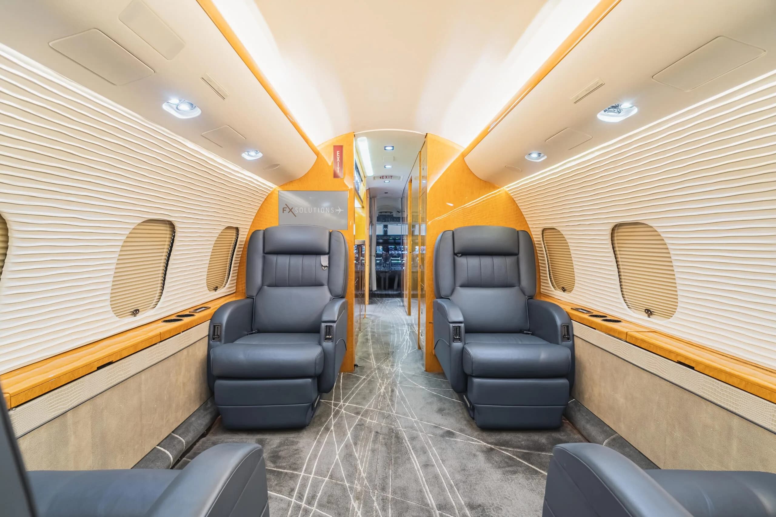 The interior of a business jet cabin, featuring two grey leather seats facing forward, light wood accents, and a view towards the galley area.