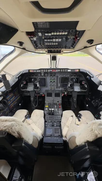 The cockpit of a business jet, showing the instrument panel, overhead panel, and two pilot seats with sheepskin covers.