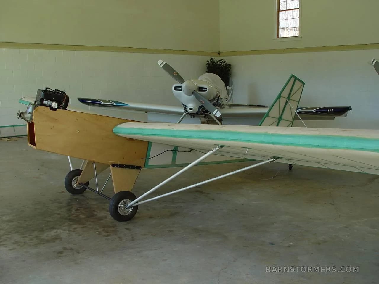 A three-quarter front view of a light aircraft with a wooden fuselage and green trim, parked in a hangar.