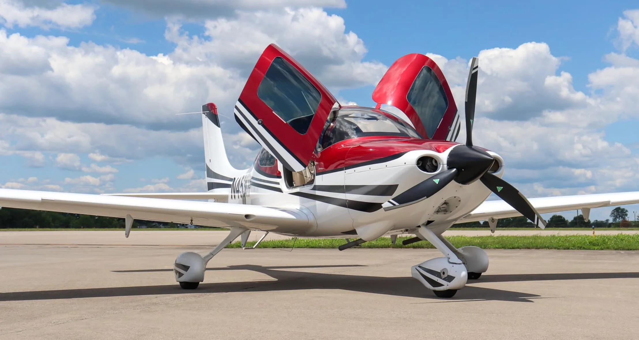 A white and red single-engine aircraft with open gull-wing doors is parked on an airfield.