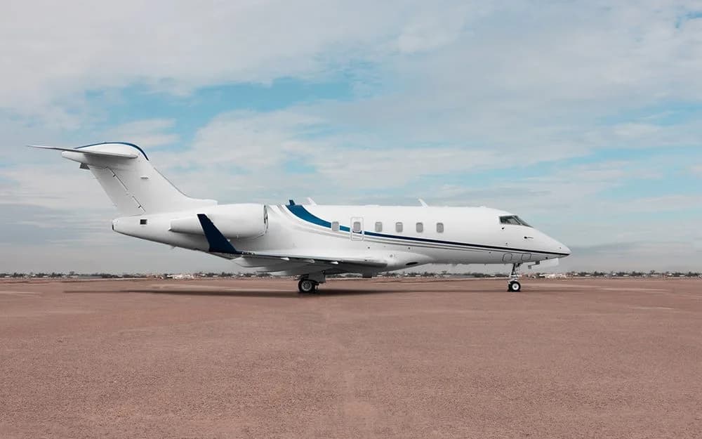 A white private jet with blue stripes is parked on an empty tarmac under a light blue sky.