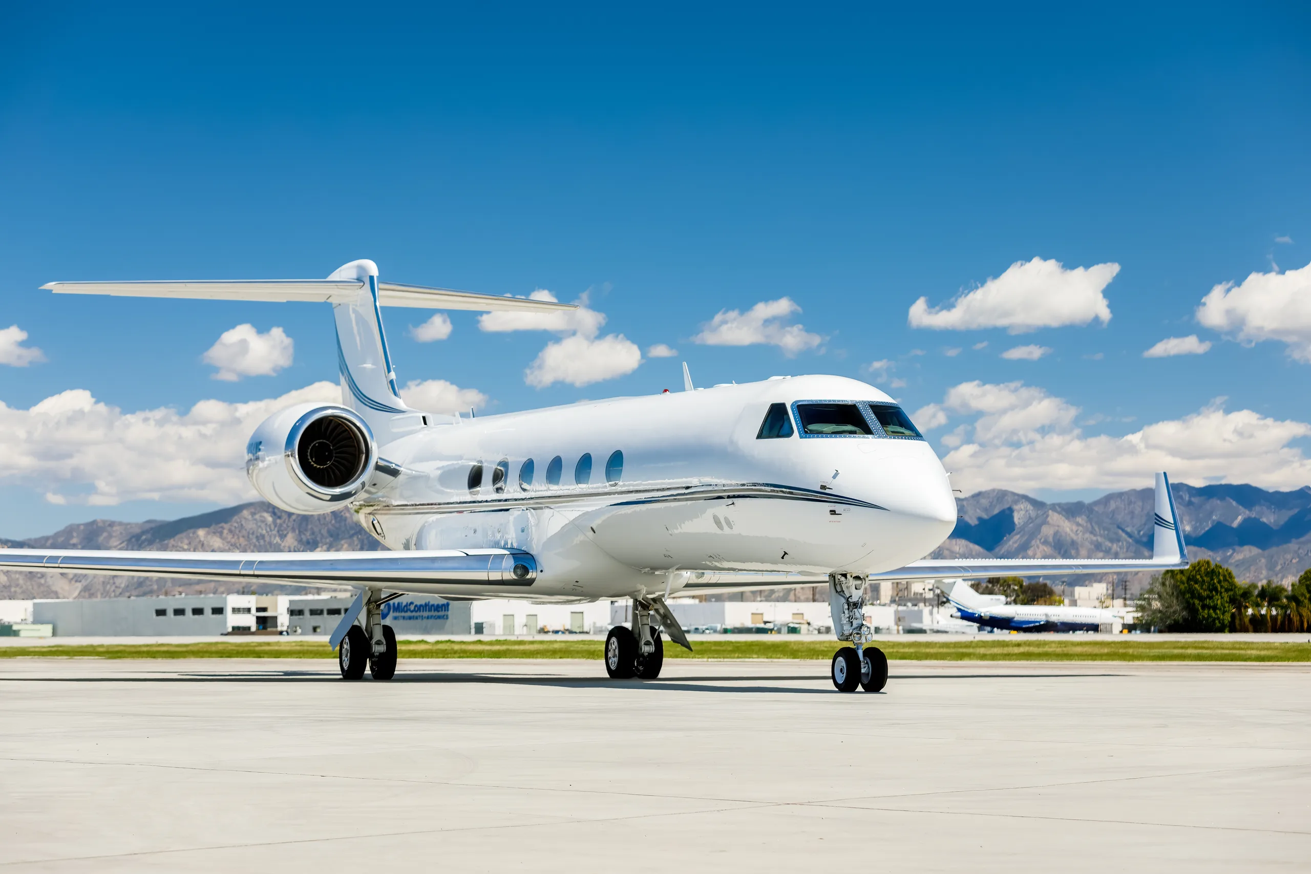A white Gulfstream G550 business jet is parked on a tarmac with mountains in the background under a blue sky.