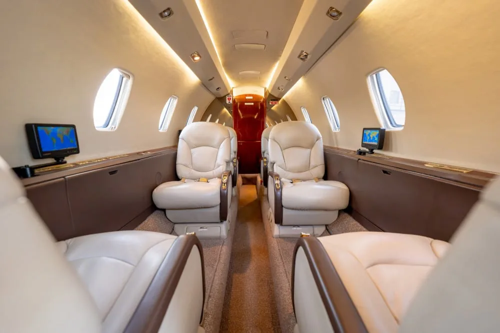 The luxurious interior cabin of a private jet, featuring beige leather seats, individual monitors, and overhead lighting.