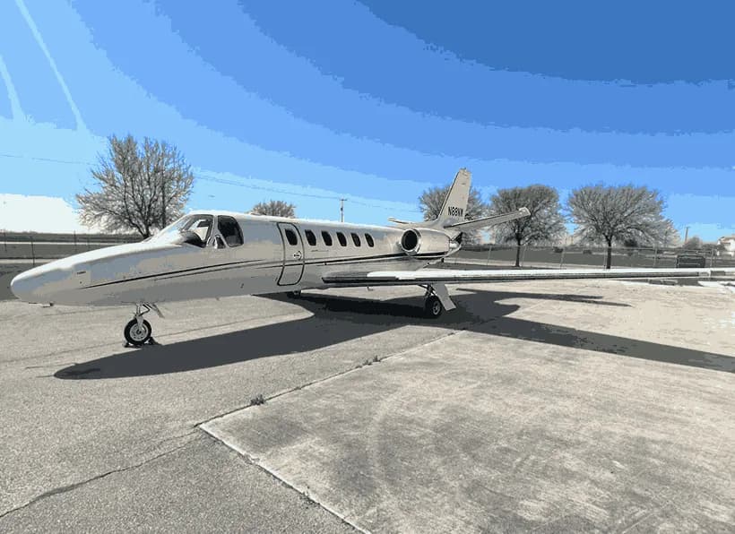 A white Cessna Citation business jet with a dark stripe is parked on a concrete ramp on a sunny day.