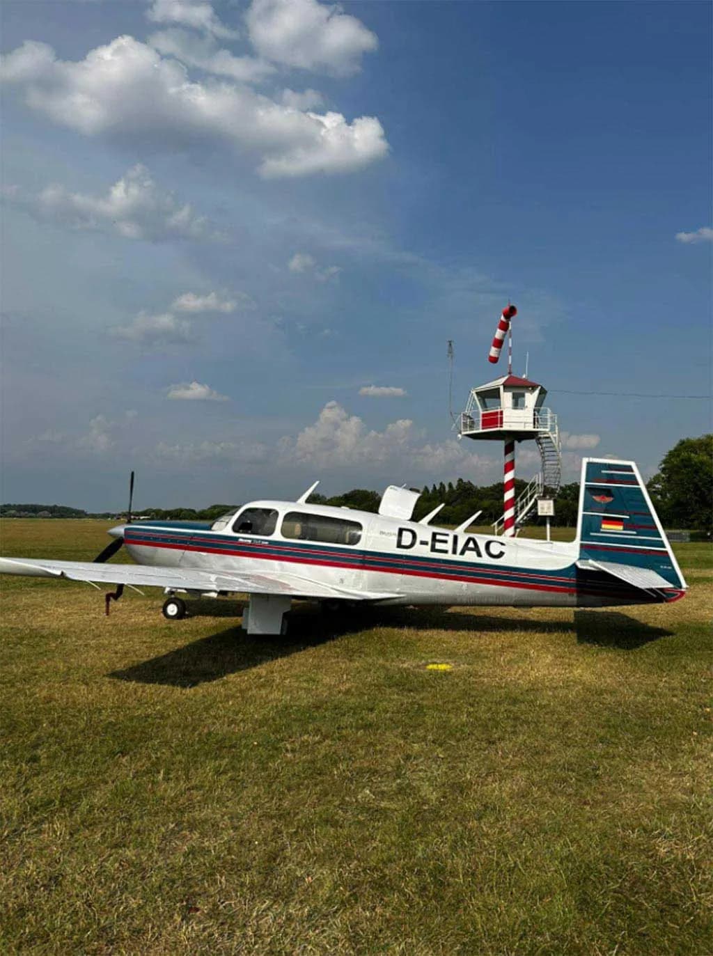 A Mooney M20J aircraft with a silver, blue, and red paint scheme parked on a grassy field next to a small red and white striped control tower.
