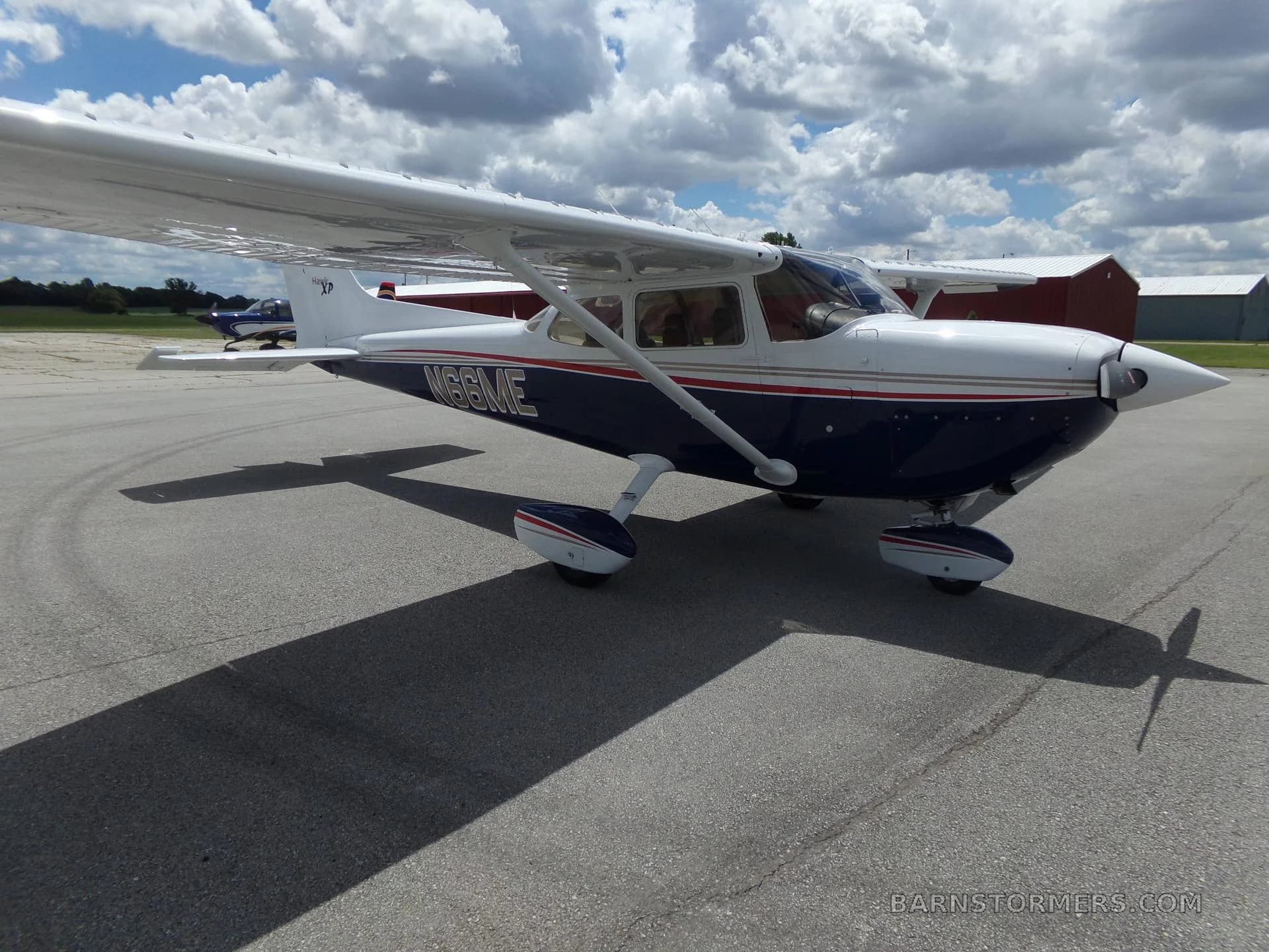 A white and blue Cessna 172 Hawk XP with red and gold stripes, seen from the front-right on an airfield with hangars in the background.