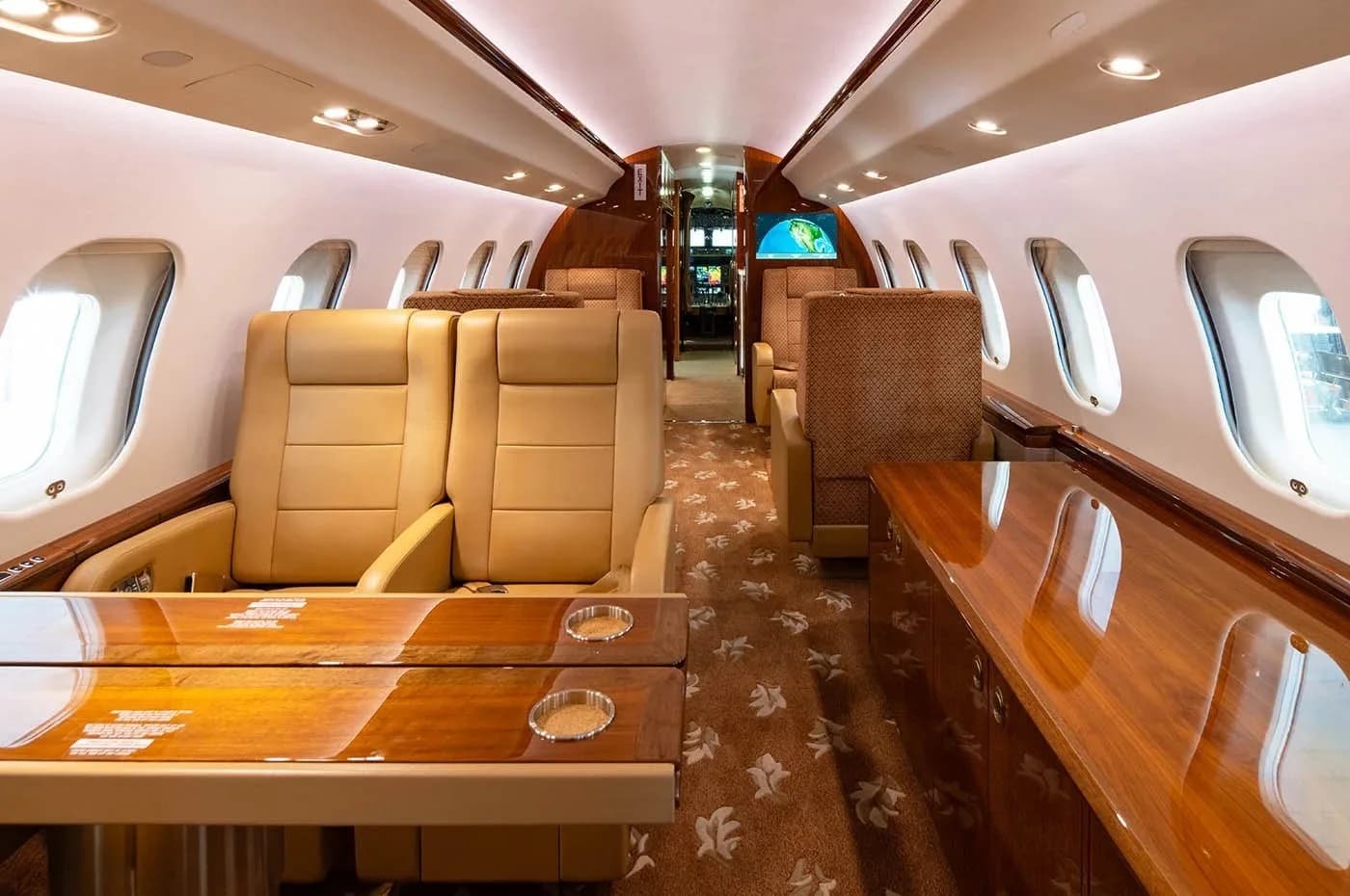 The luxurious aircraft cabin features tan leather seats, wooden tables, and a patterned carpet.