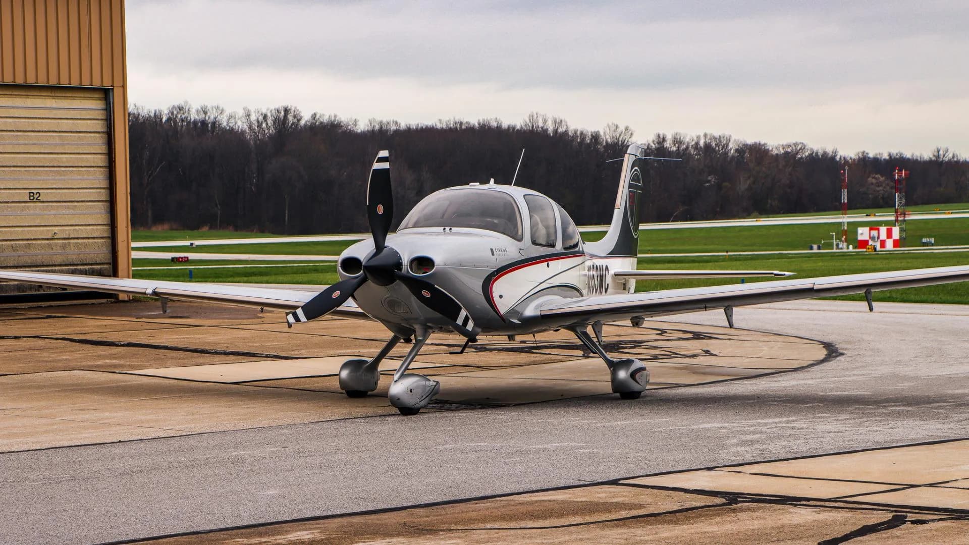 A silver and white Cirrus SR22 aircraft with N553MC registration is parked on an airport tarmac next to a hangar.