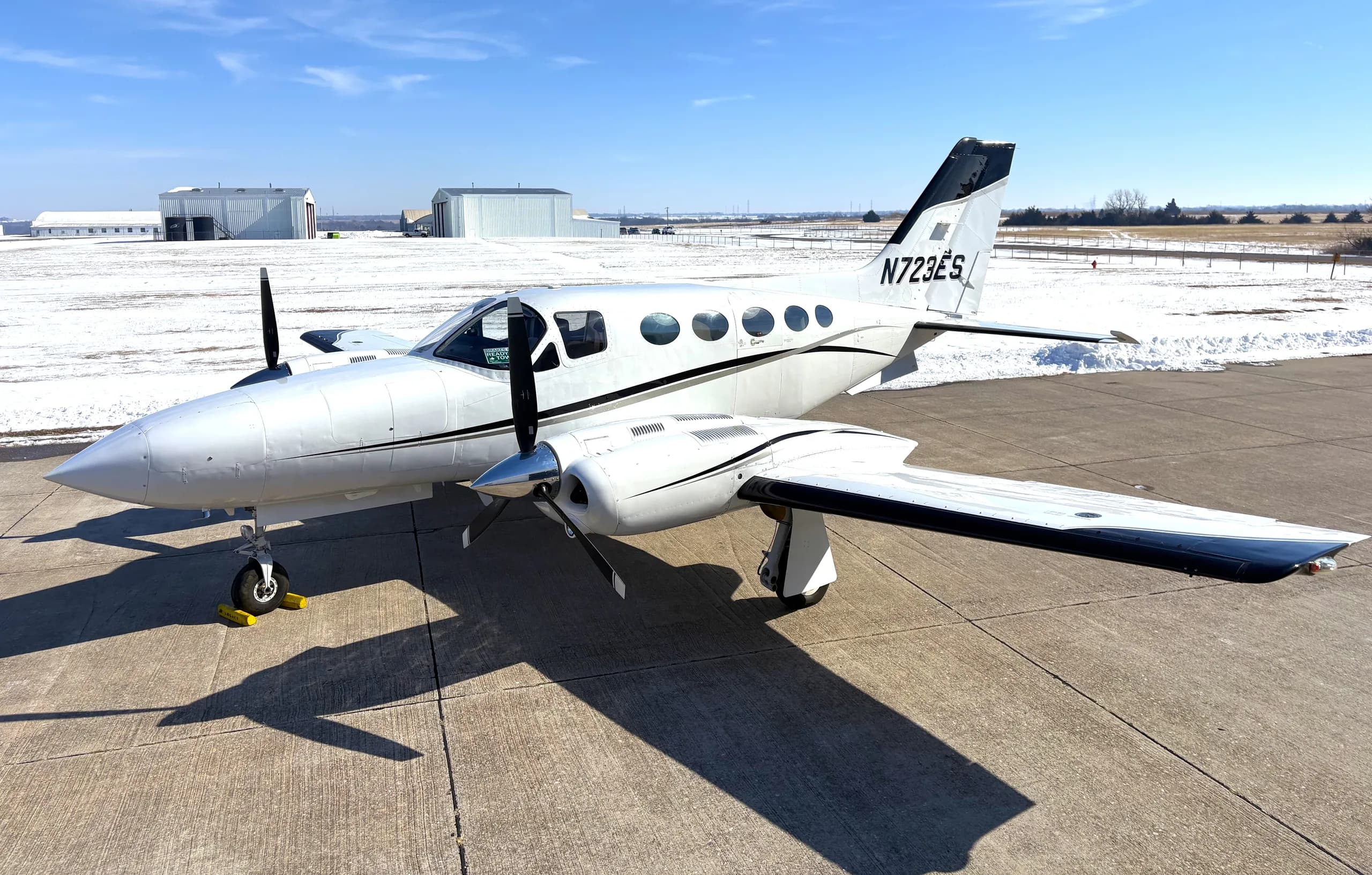 A white twin-engine propeller aircraft, N723ES, with black stripes, parked on a snowy airfield.