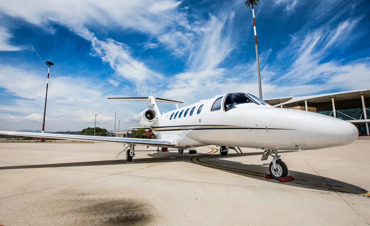 A white Cessna CitationJet with black and gold stripes, registration N420CJ, parked on an airport tarmac under a blue sky with wispy clouds.