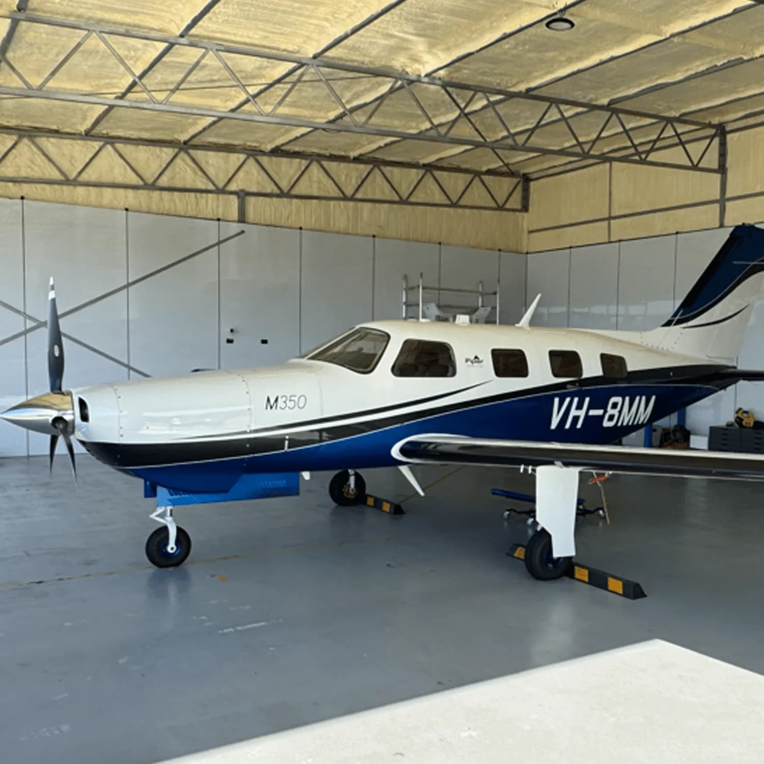 A white and blue Piper M350 aircraft with registration VH-8MM parked inside a hangar.
