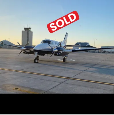 A silver and dark blue twin-engine turboprop aircraft, likely a King Air, parked on an airport tarmac with an air traffic control tower in the background.