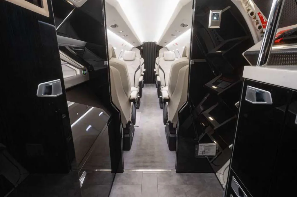 A view down the aisle of a private jet's interior cabin, featuring white seats and dark, reflective side panels.