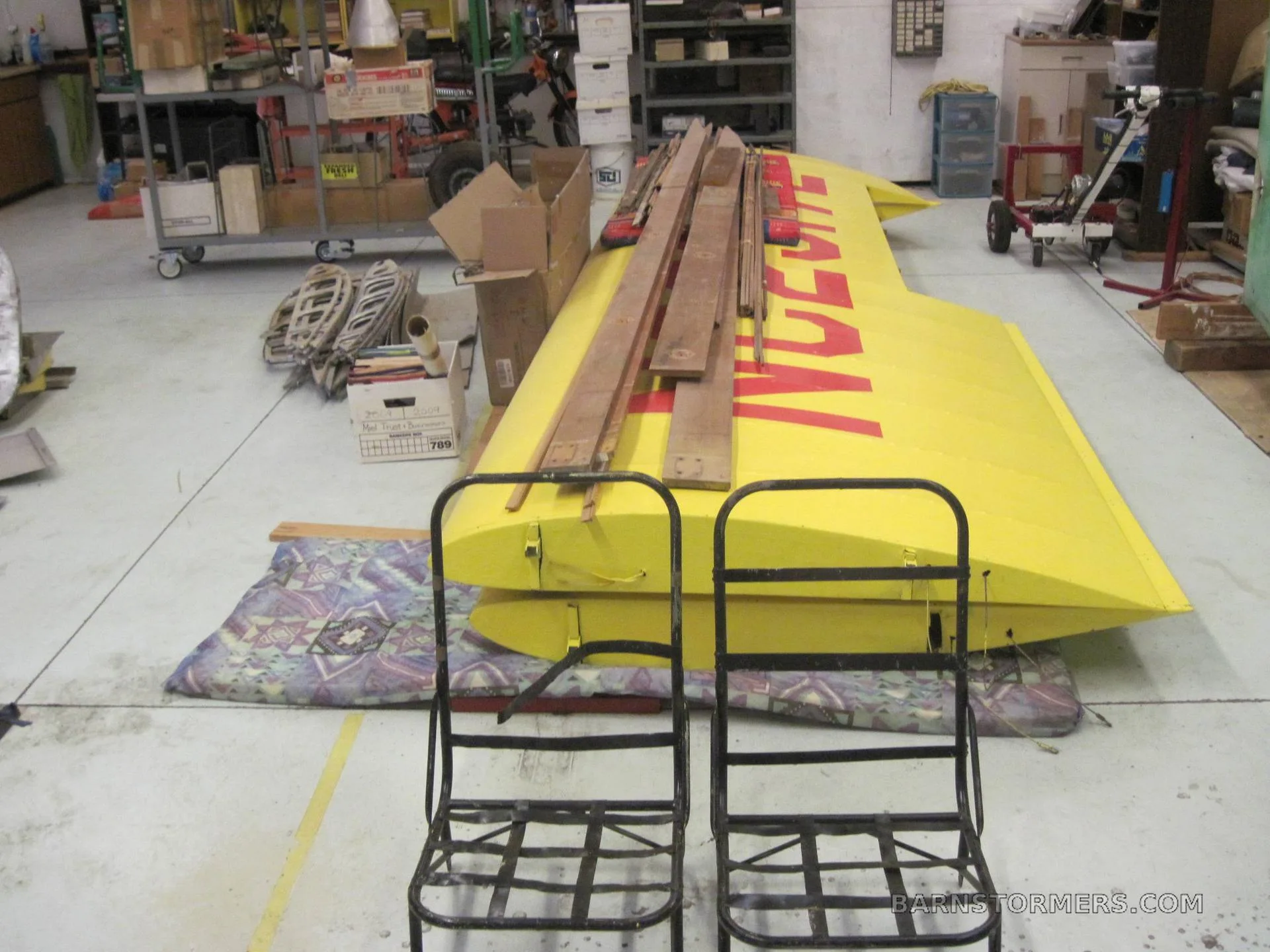 A large yellow aircraft wing with red lettering, resting on cushions in a workshop, with two metal seats in the foreground.