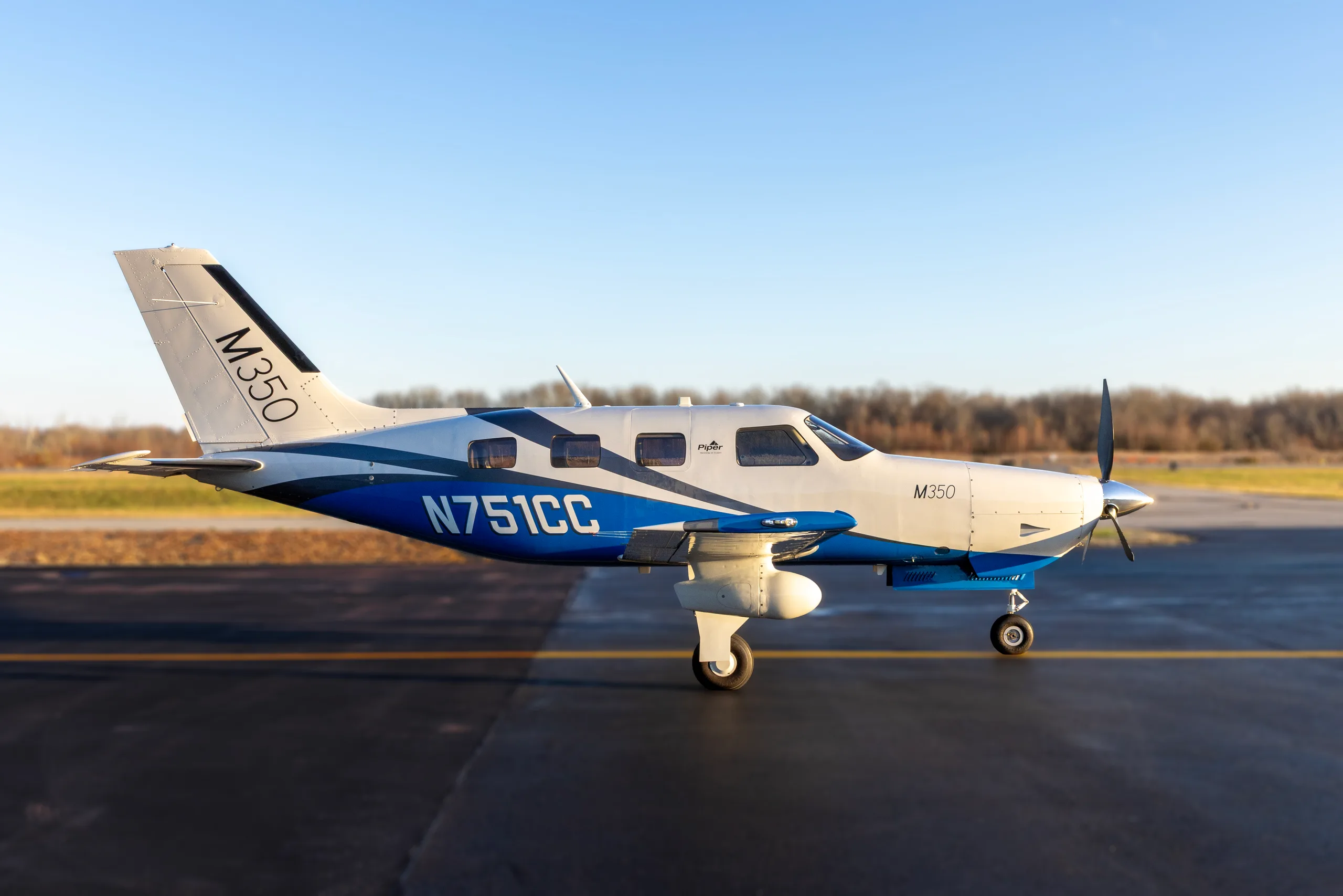 A Piper M350 aircraft, registration N751CC, with a white and blue paint scheme, parked on an airfield at sunset.