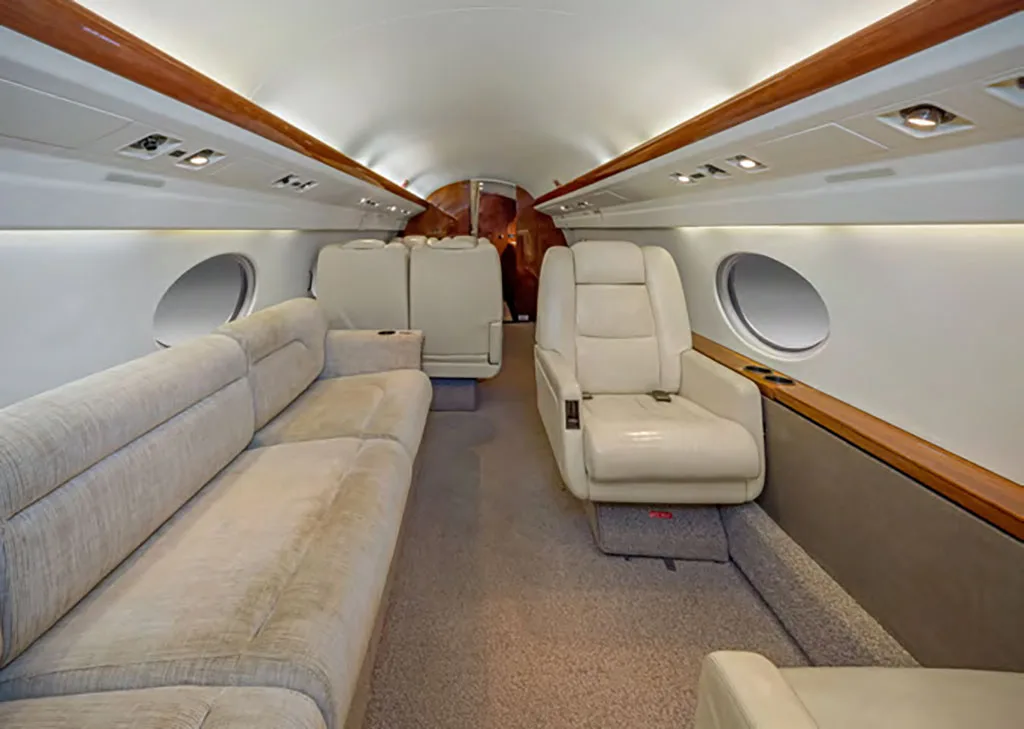 The interior cabin of a private jet shows a beige sofa on one side and a single beige leather seat on the other, with wood trim.