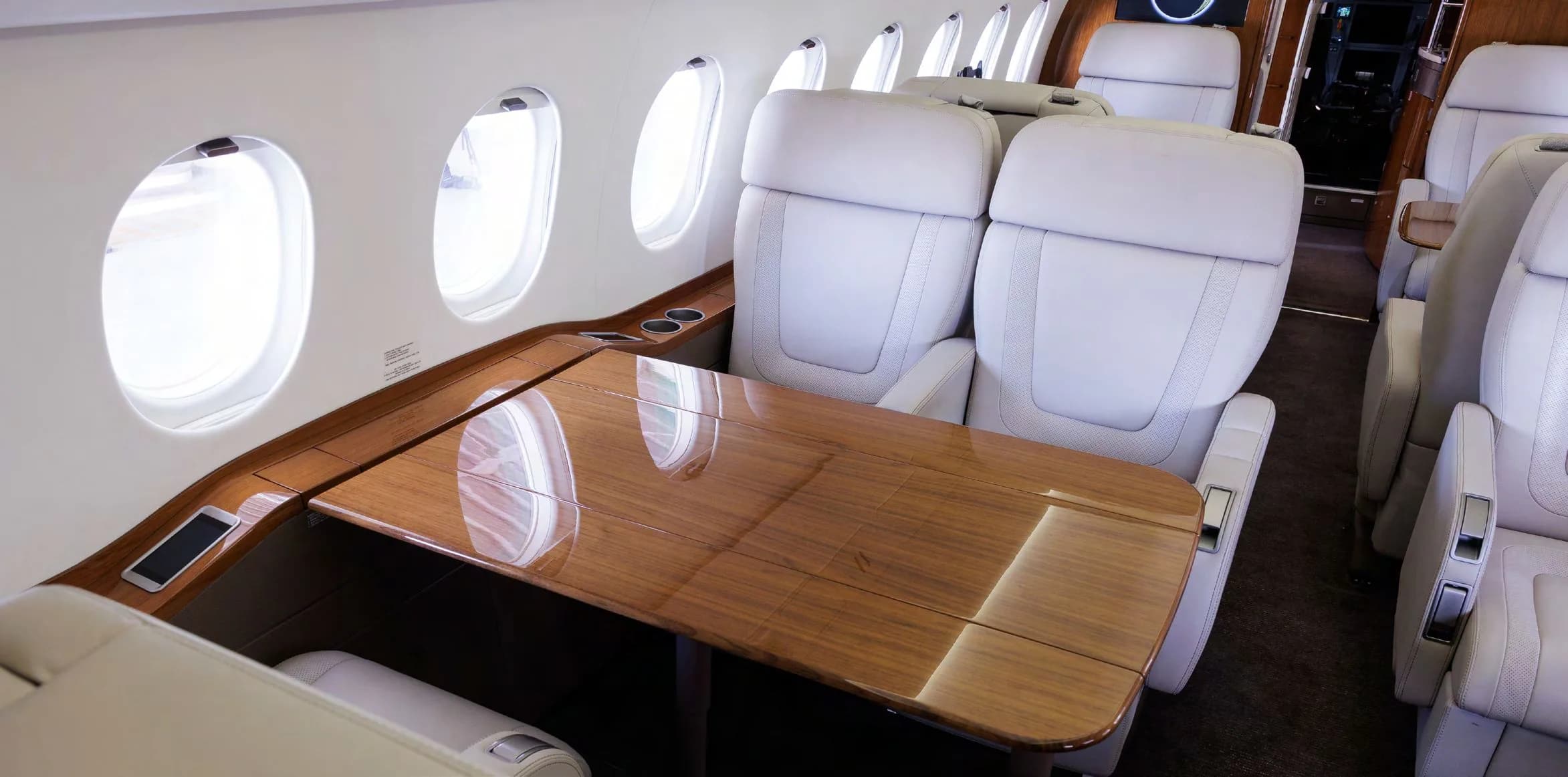 A close-up view of the business jet's cabin showing light grey seats, a large polished wooden table, and windows.