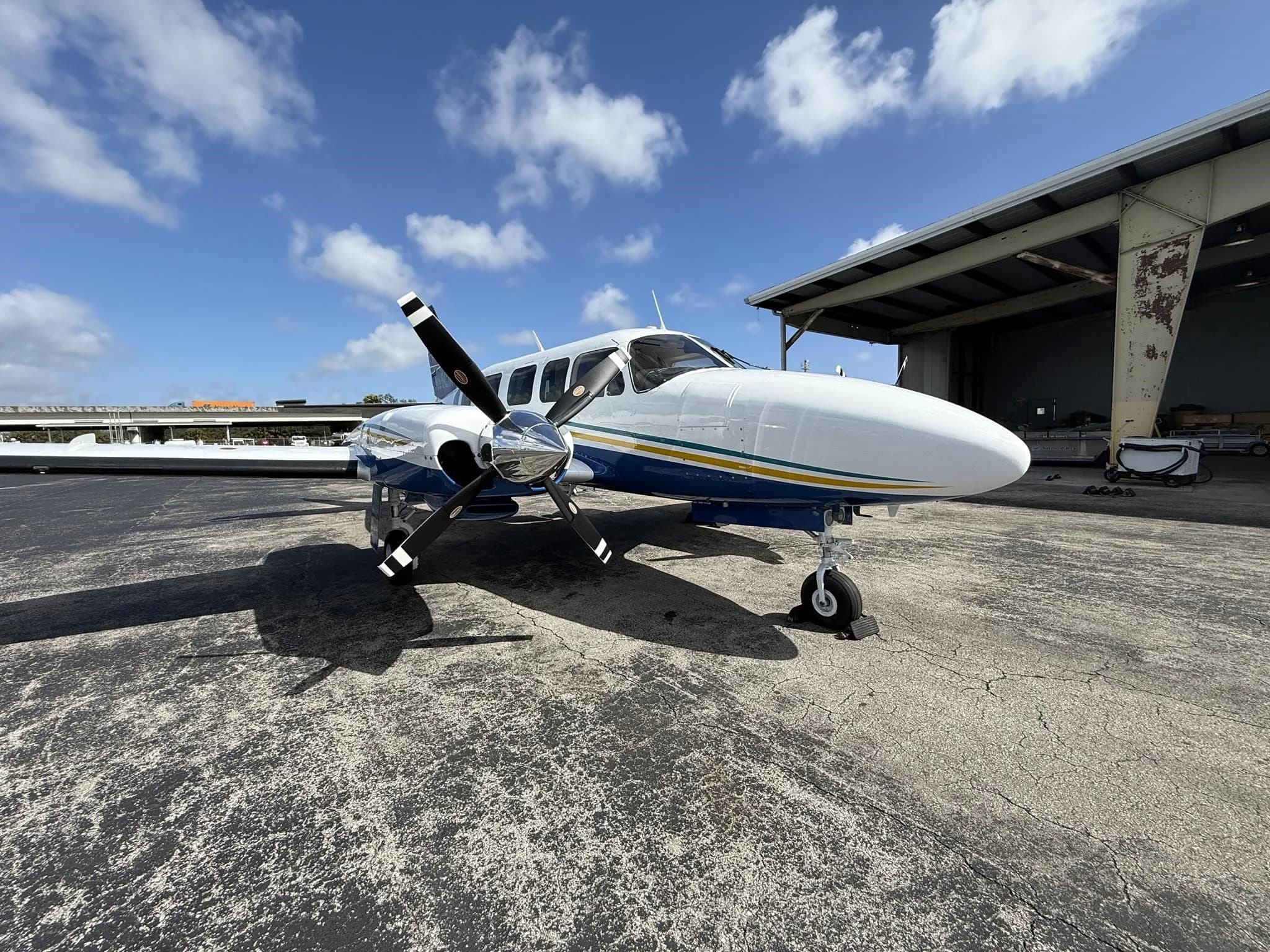 A white and blue twin-engine propeller aircraft with yellow and green stripes, parked on an airport tarmac next to a hangar.