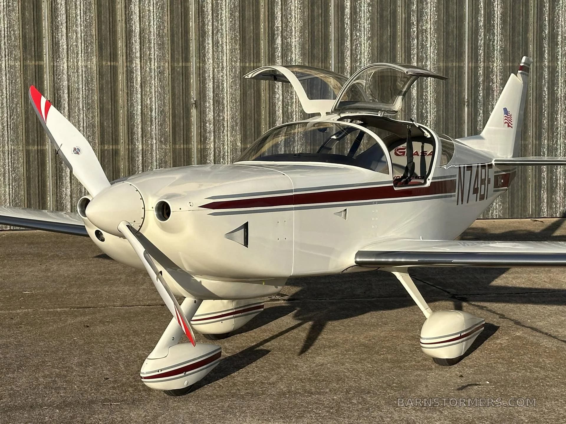 A white single-engine aircraft with a maroon stripe and registration N74BF is parked on concrete with its canopy open, viewed from the front.