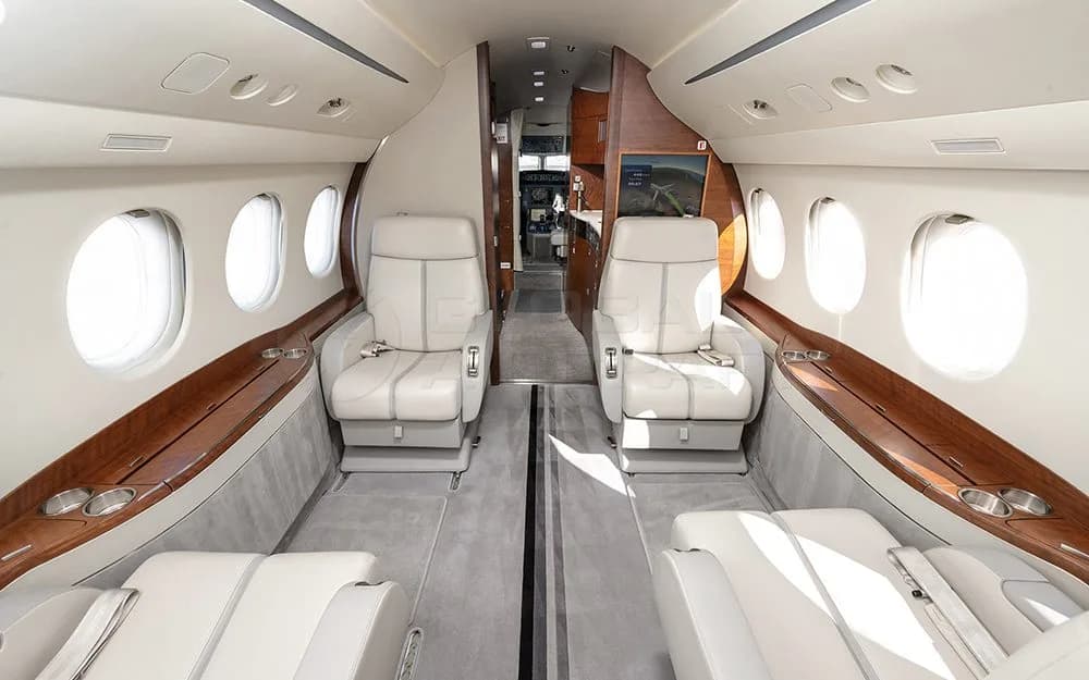 A view down the aisle of a private jet's cabin, showing rows of light-colored leather seats and a screen at the far end.