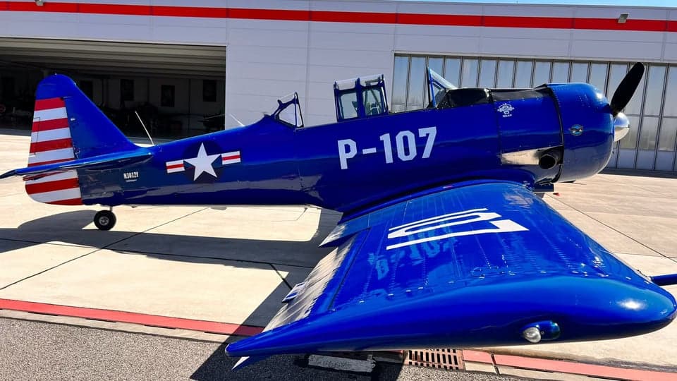 A blue North American Aviation T-6 Texan aircraft parked in front of a hangar with a white star and red and white stripes on the tail.