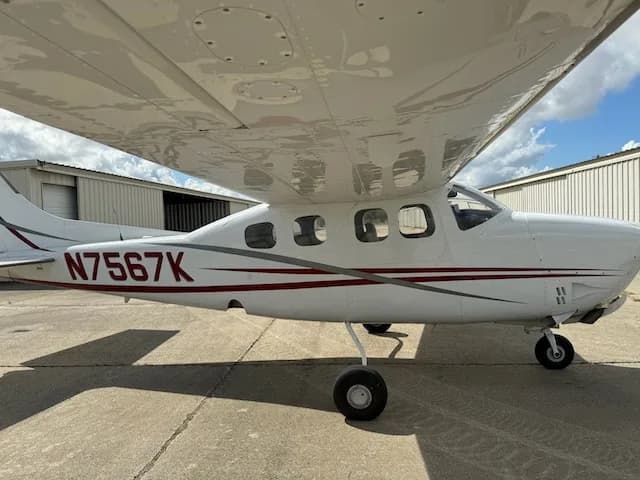 A side view of a white Cessna 206 with red and grey stripes, showing the tail number N7567K, parked near hangars.