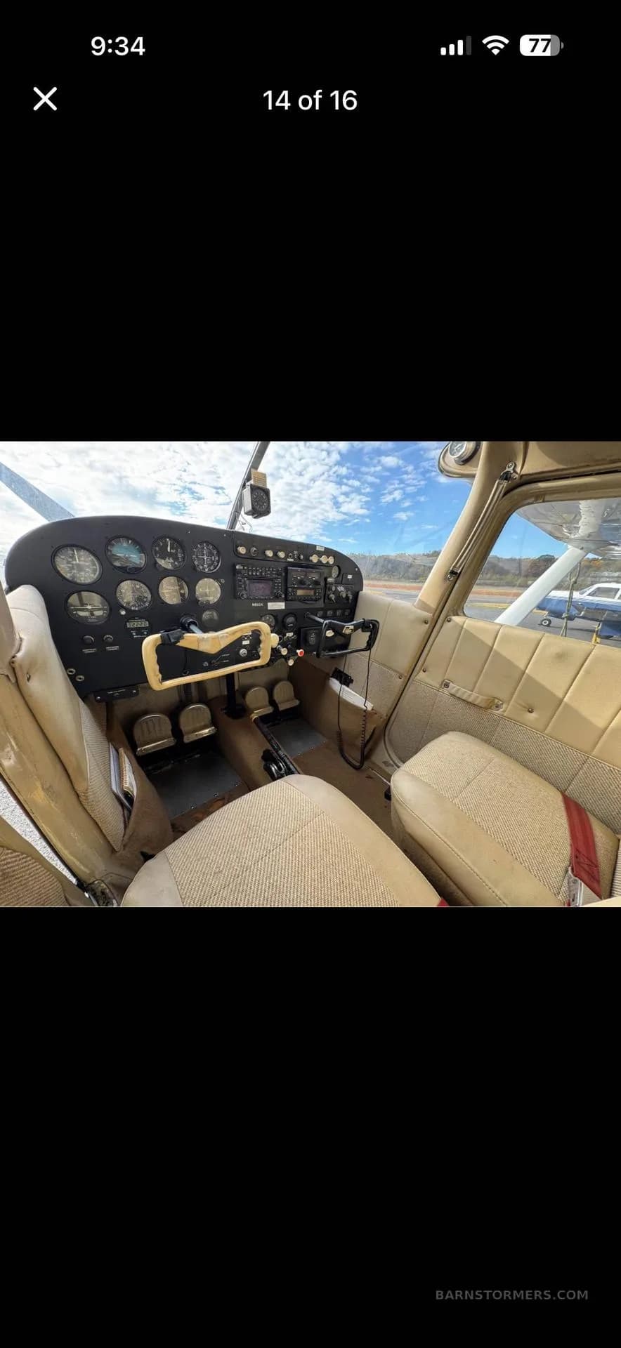 The cockpit of a light aircraft features a beige interior, a black instrument panel, and a view of the sky outside.