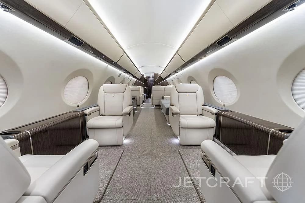 The luxurious interior cabin of a private jet, featuring light cream leather seats and a central aisle with soft lighting.