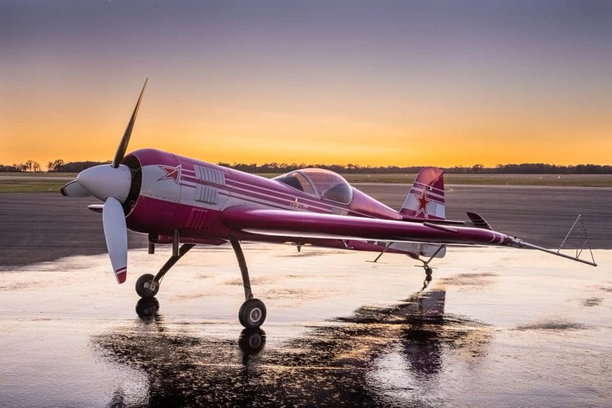 A pink and white Sukhoi Su-26 aircraft parked on a wet tarmac at sunset.