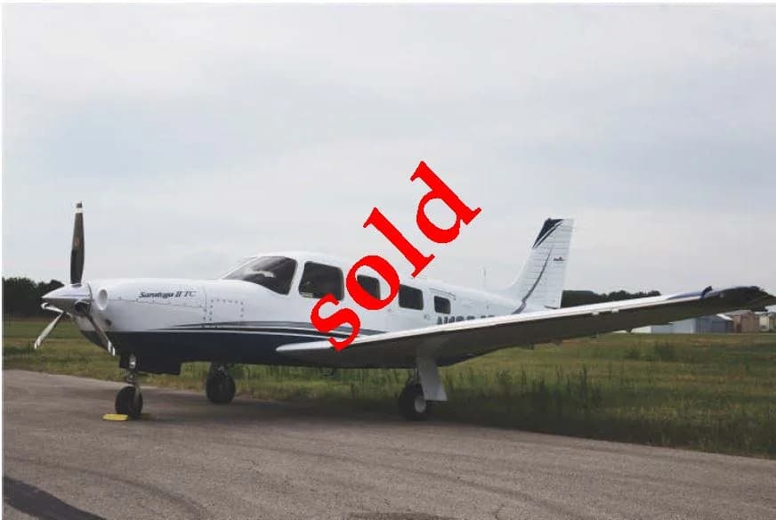 A white and blue Piper Saratoga II TC single-engine aircraft with "SOLD" text, parked on an airfield.