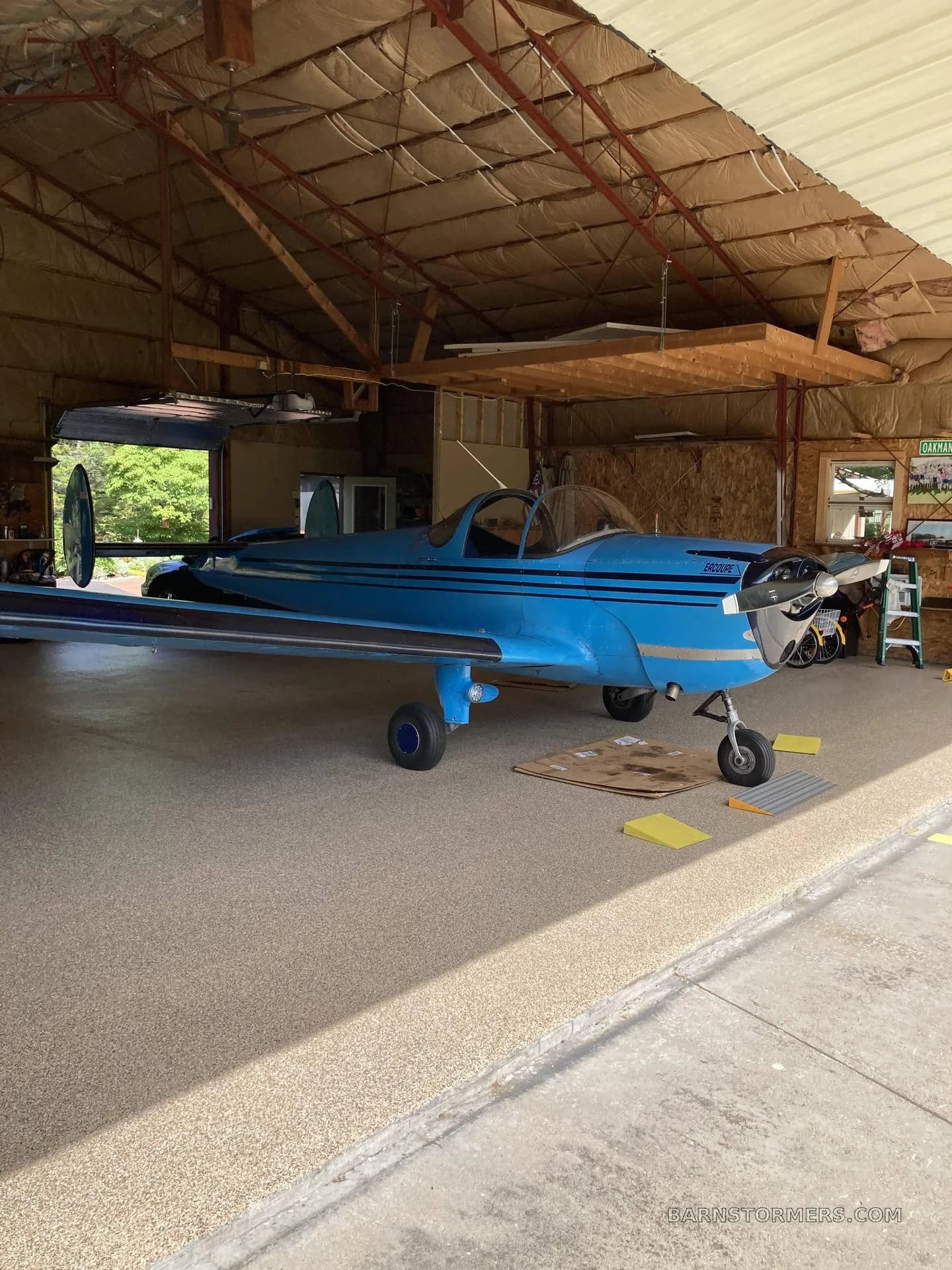 A bright blue Ercoupe aircraft with black stripes parked inside a hangar with a speckled floor.