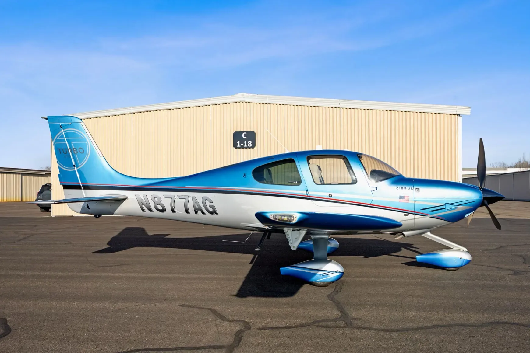 A blue and silver Cirrus SR22T aircraft, registration N877AG, parked on an airfield in front of a hangar.