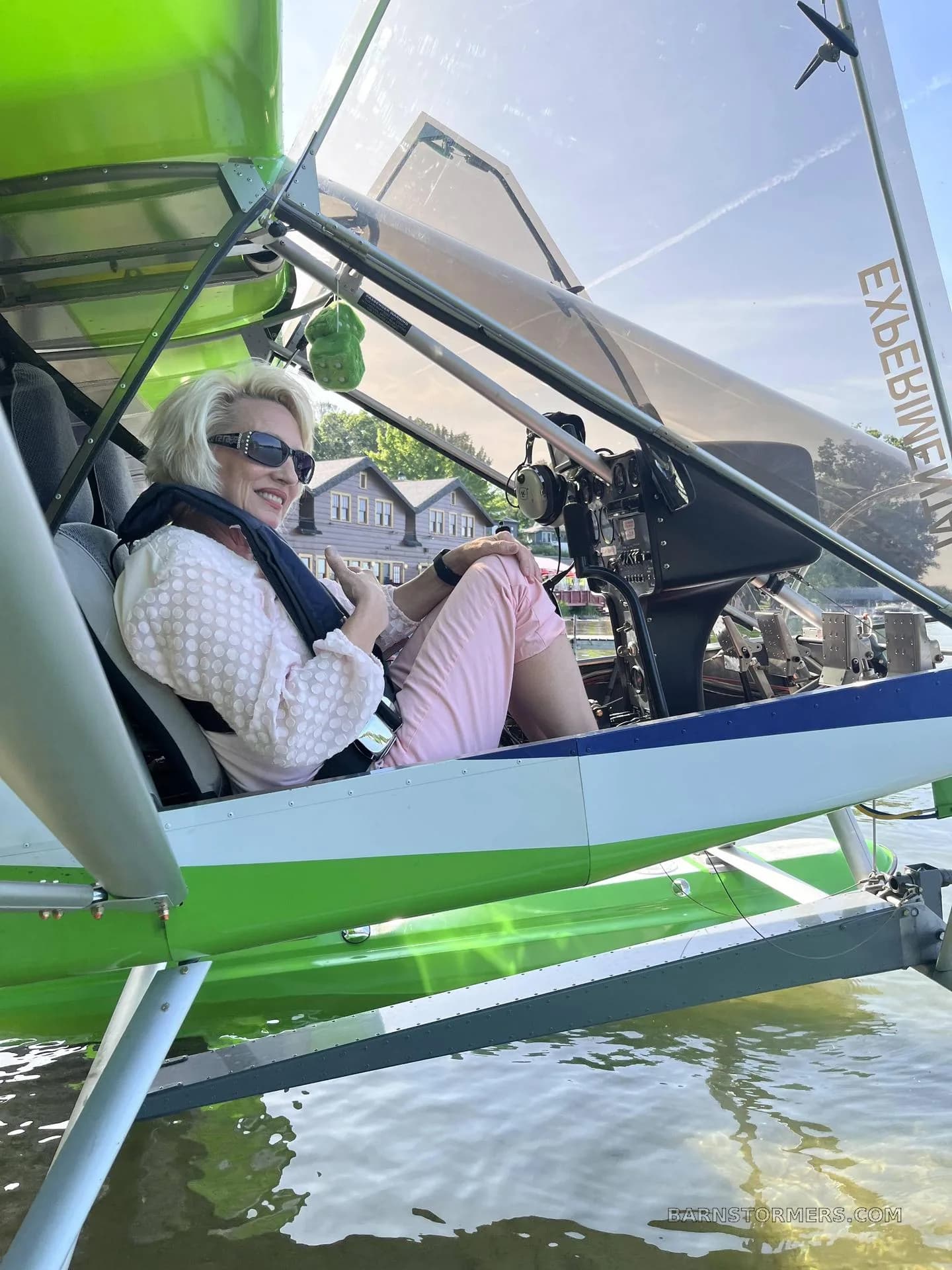 A smiling woman with blonde hair and sunglasses sits in the cockpit of a green and blue ultralight seaplane, which is partially in the water.
