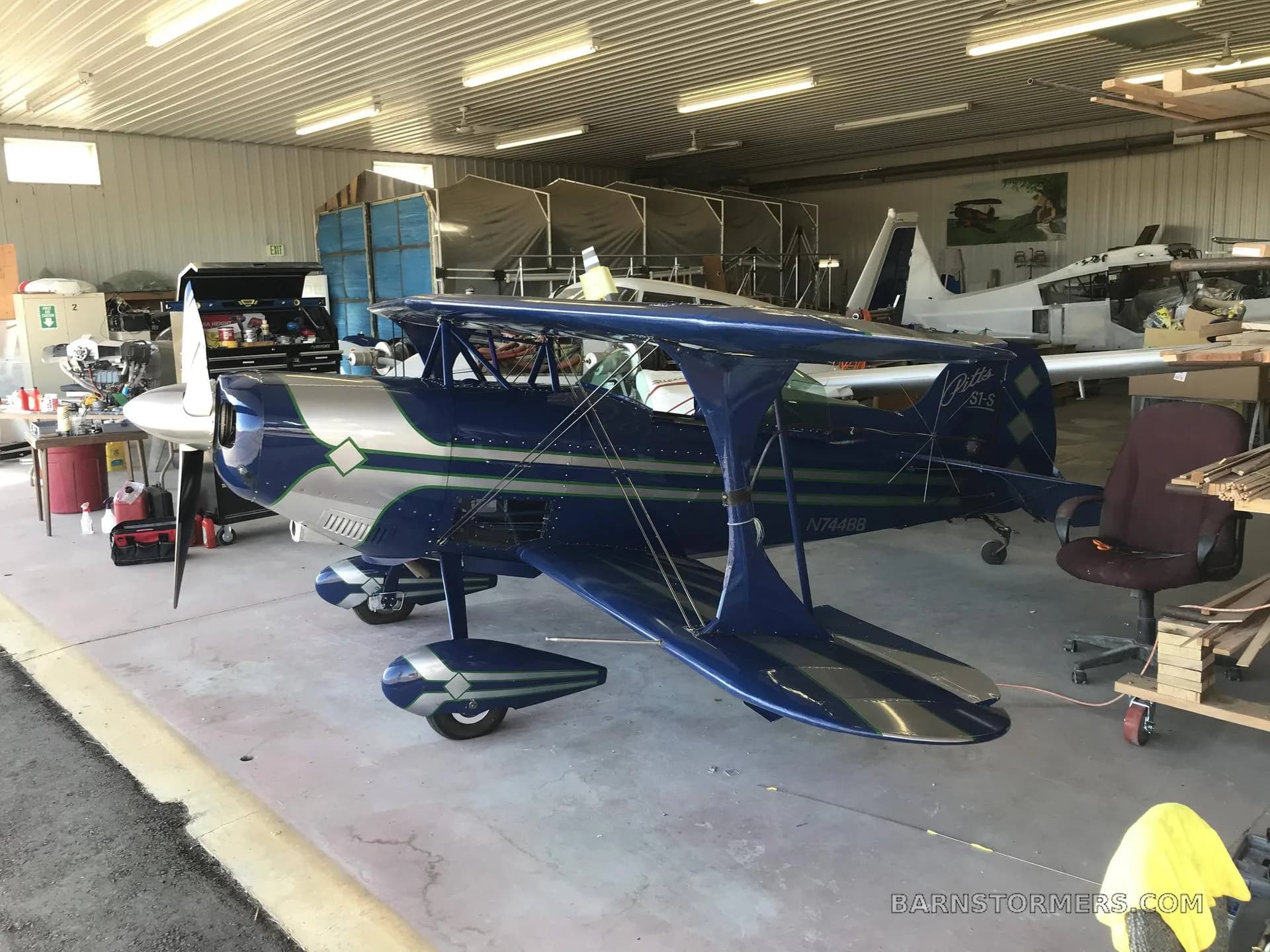 A blue and silver Pitts S1-S biplane with green stripes is parked in a hangar with other aircraft in the background.