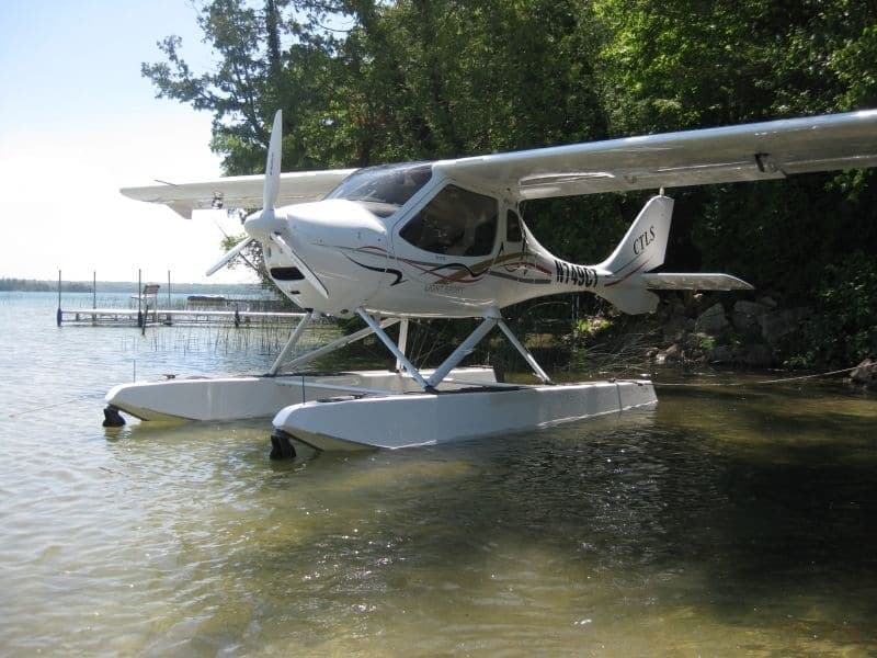 A white CTLS light sport aircraft with floats is partially submerged in a lake near a dock.