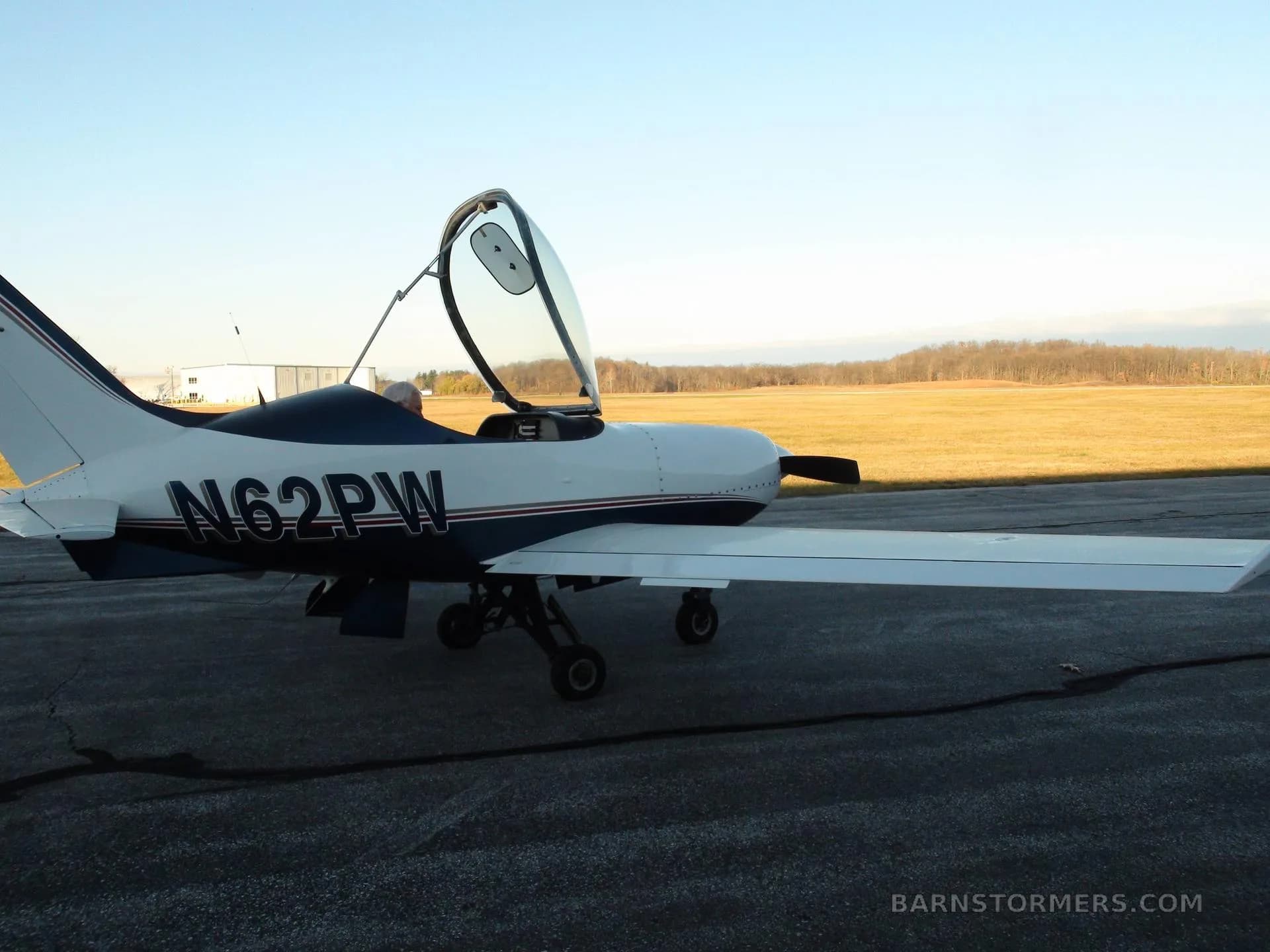 A white and blue single-engine aircraft with its canopy open, parked on an asphalt runway with a field in the background.