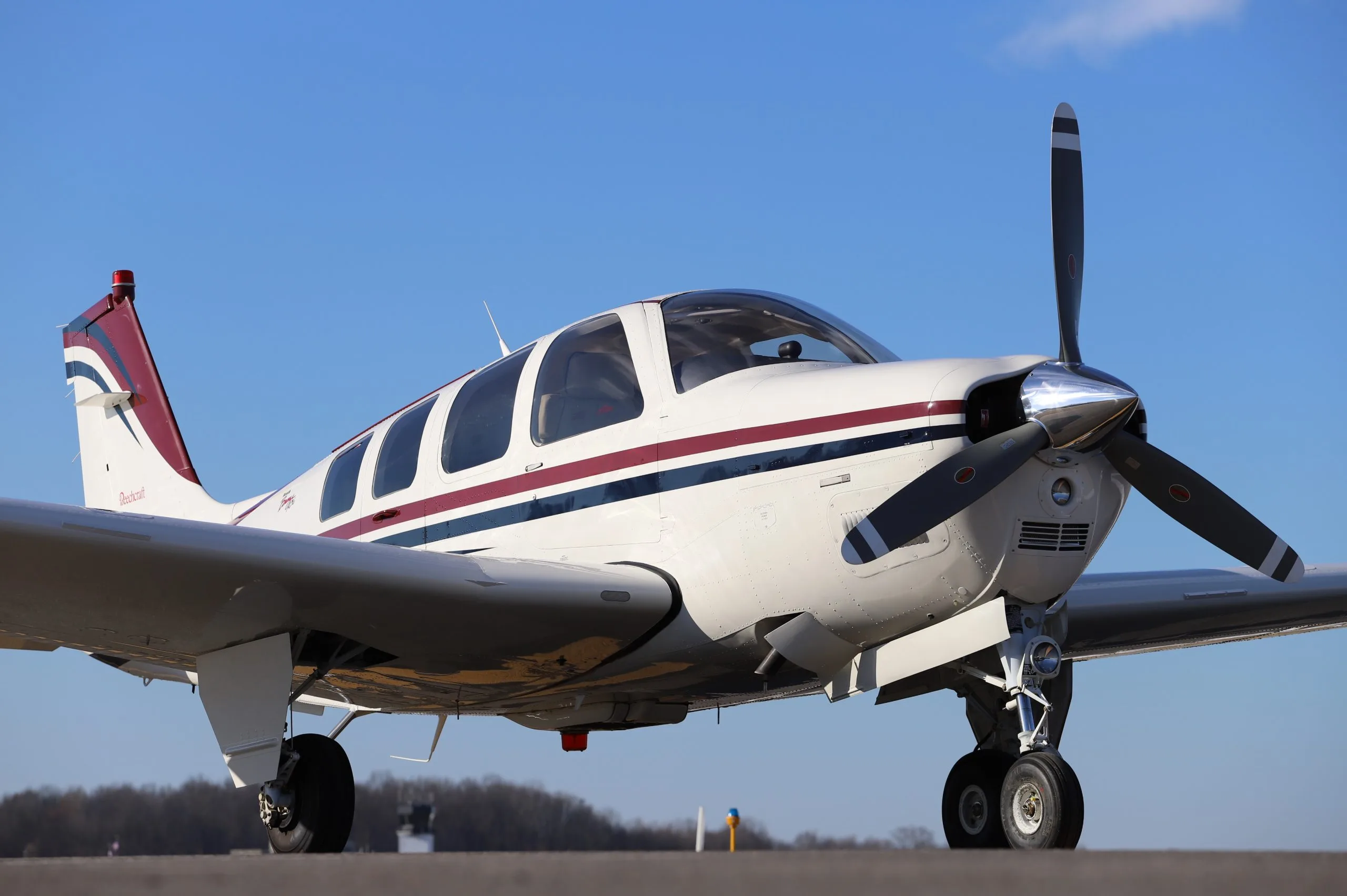 A white Beechcraft Bonanza with maroon and blue stripes is parked on the tarmac under a clear blue sky.