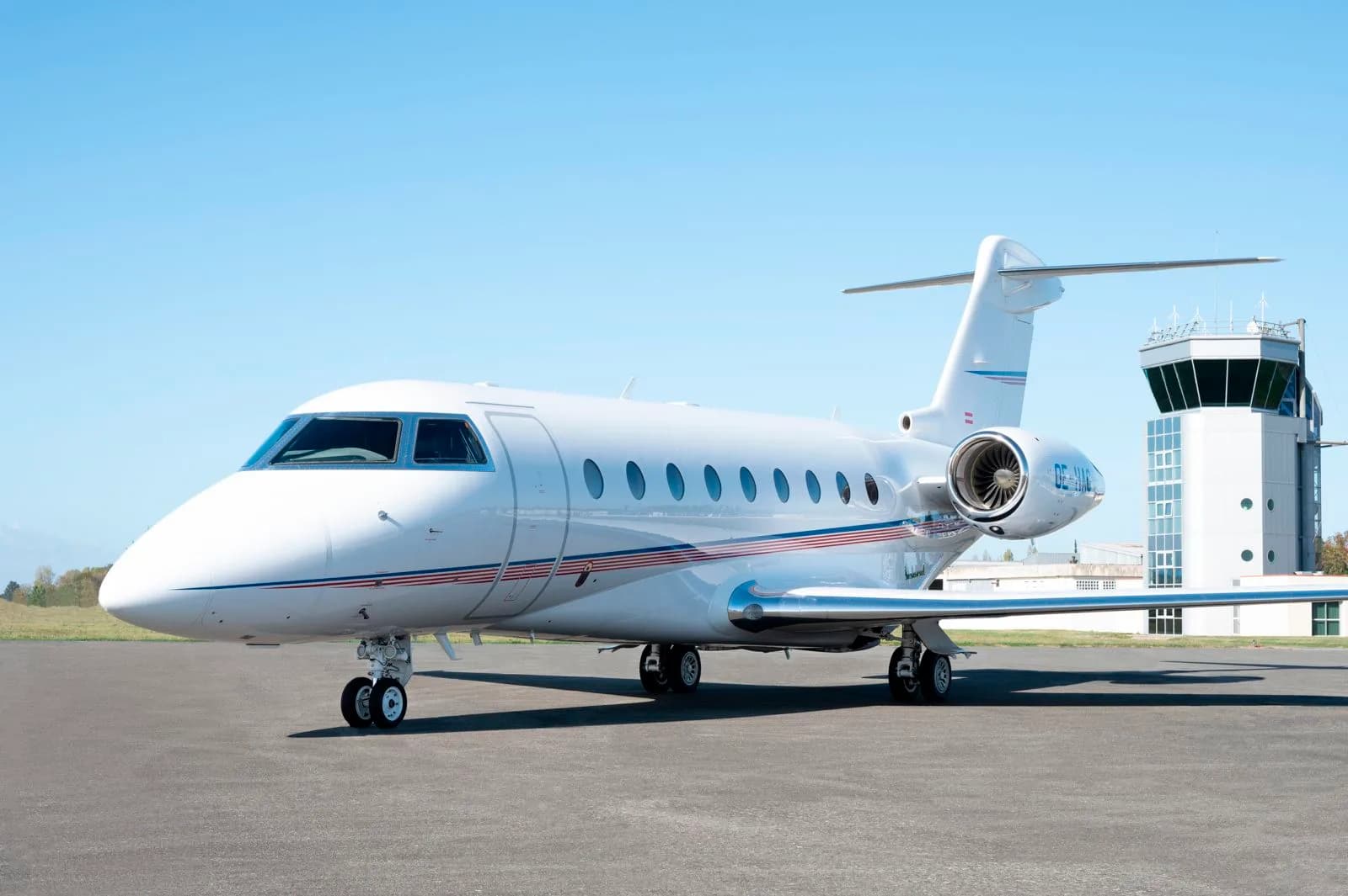 A white business jet with red and blue stripes is parked on an airport tarmac, with an air traffic control tower in the background.