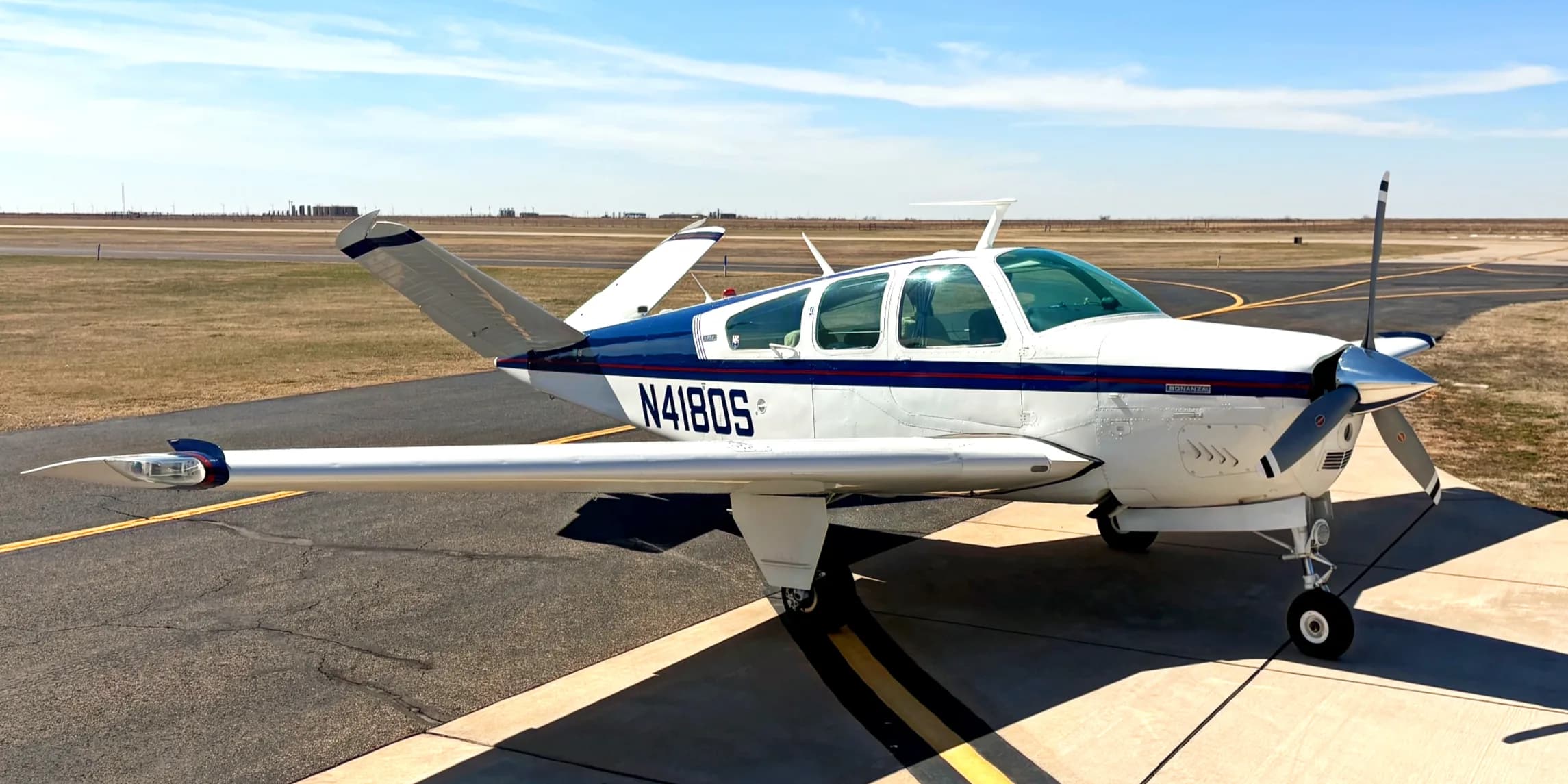 A white and blue single-engine propeller aircraft, N4180S, parked on an airfield under a clear sky.
