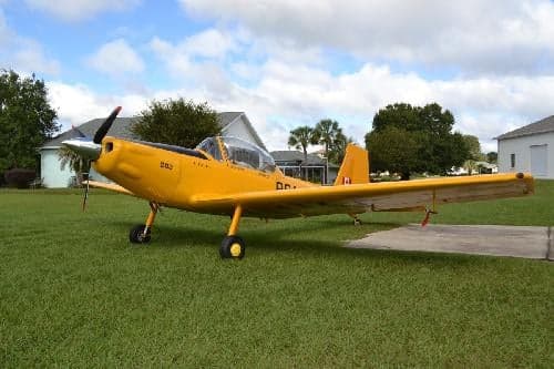A yellow aircraft is parked on a grassy lawn in front of a house on a sunny day.