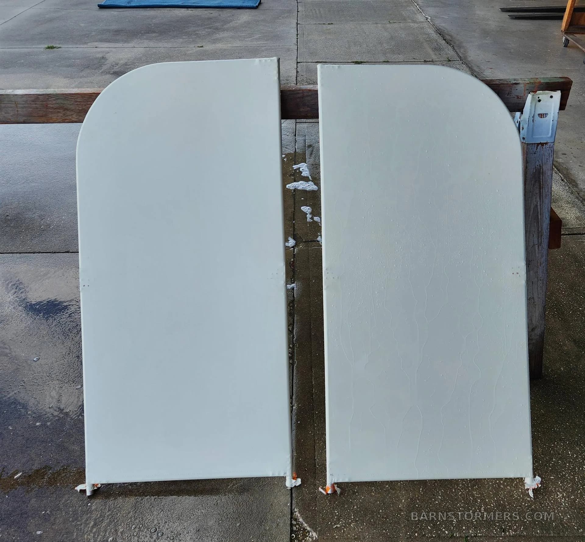 Two rounded, light grey aircraft components, possibly fairings, resting vertically on sawhorses on a wet concrete pad.