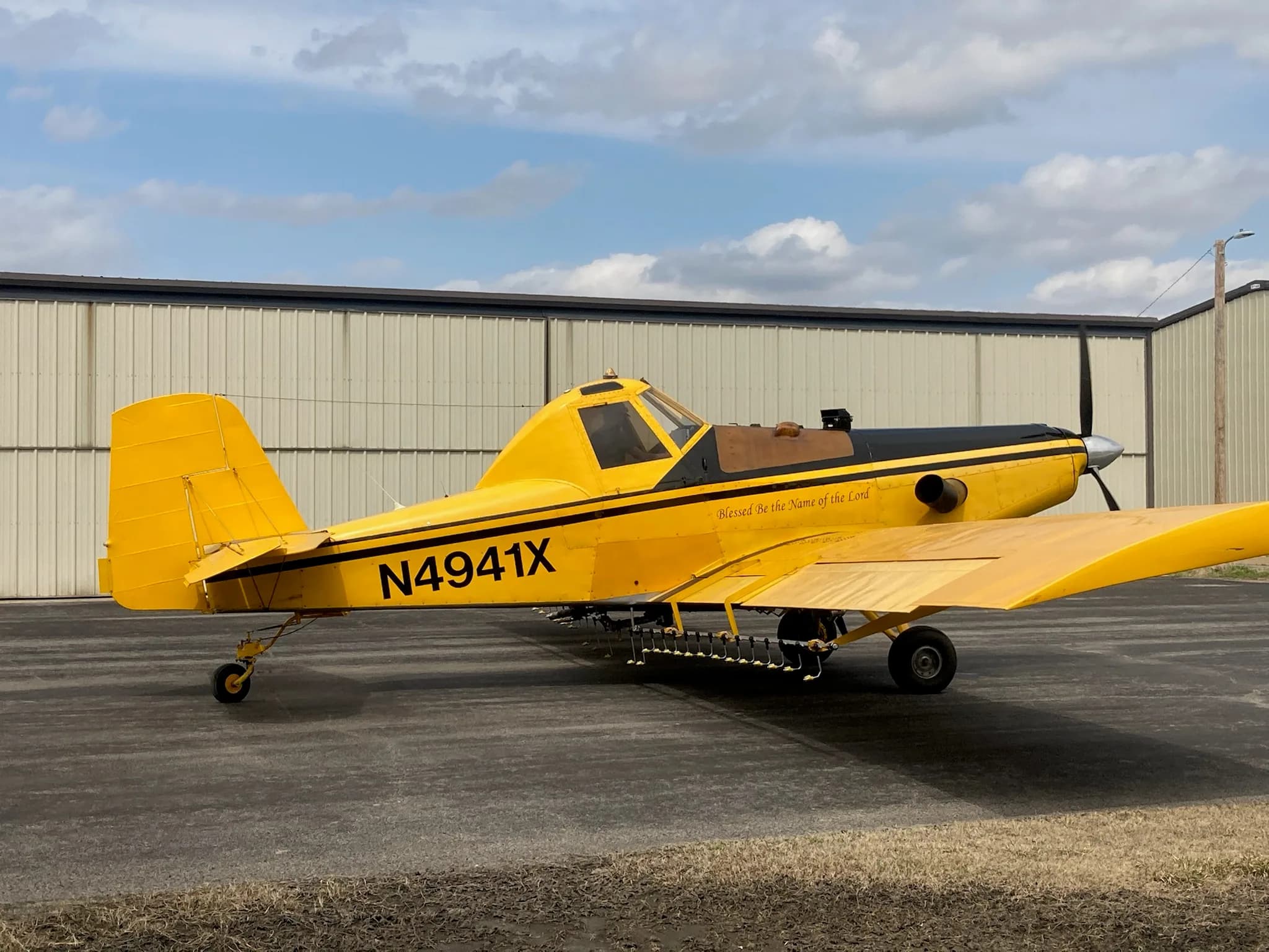 A yellow and black crop duster aircraft, N4941X, with 'Blessed be the Name of the Lord' painted on its side, parked in front of a hangar.