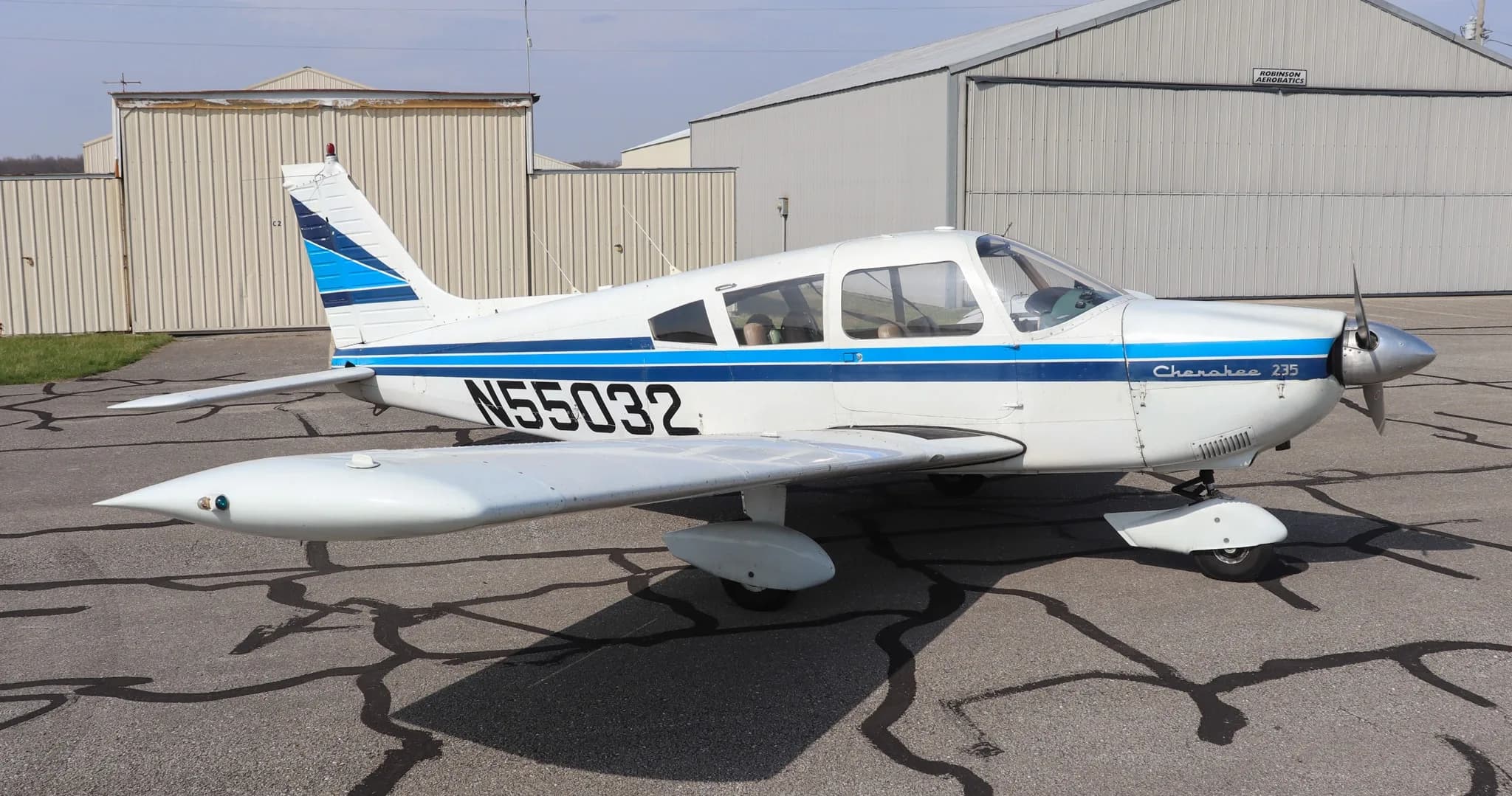 A white Piper Cherokee 235 aircraft with blue stripes and registration N55032 parked on an asphalt ramp next to hangars.