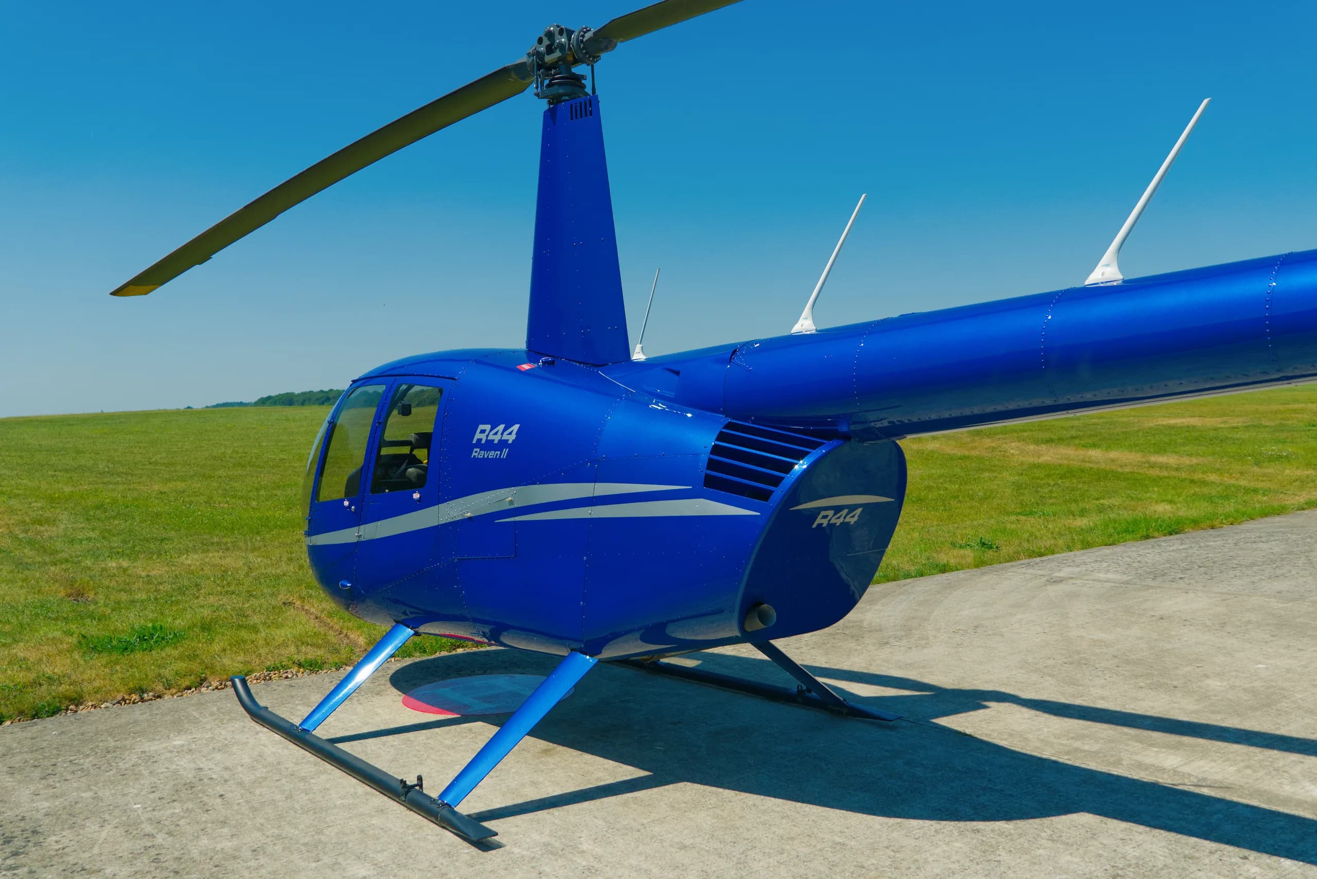 A blue Robinson R44 Raven II helicopter with a white stripe sits on a concrete pad next to a grassy field under a clear sky.