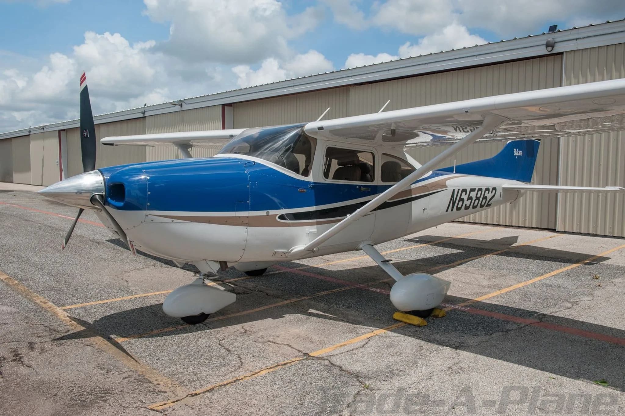 A front-left view of a white and blue Cessna Skylane N65862 parked on an asphalt ramp under a partly cloudy sky.