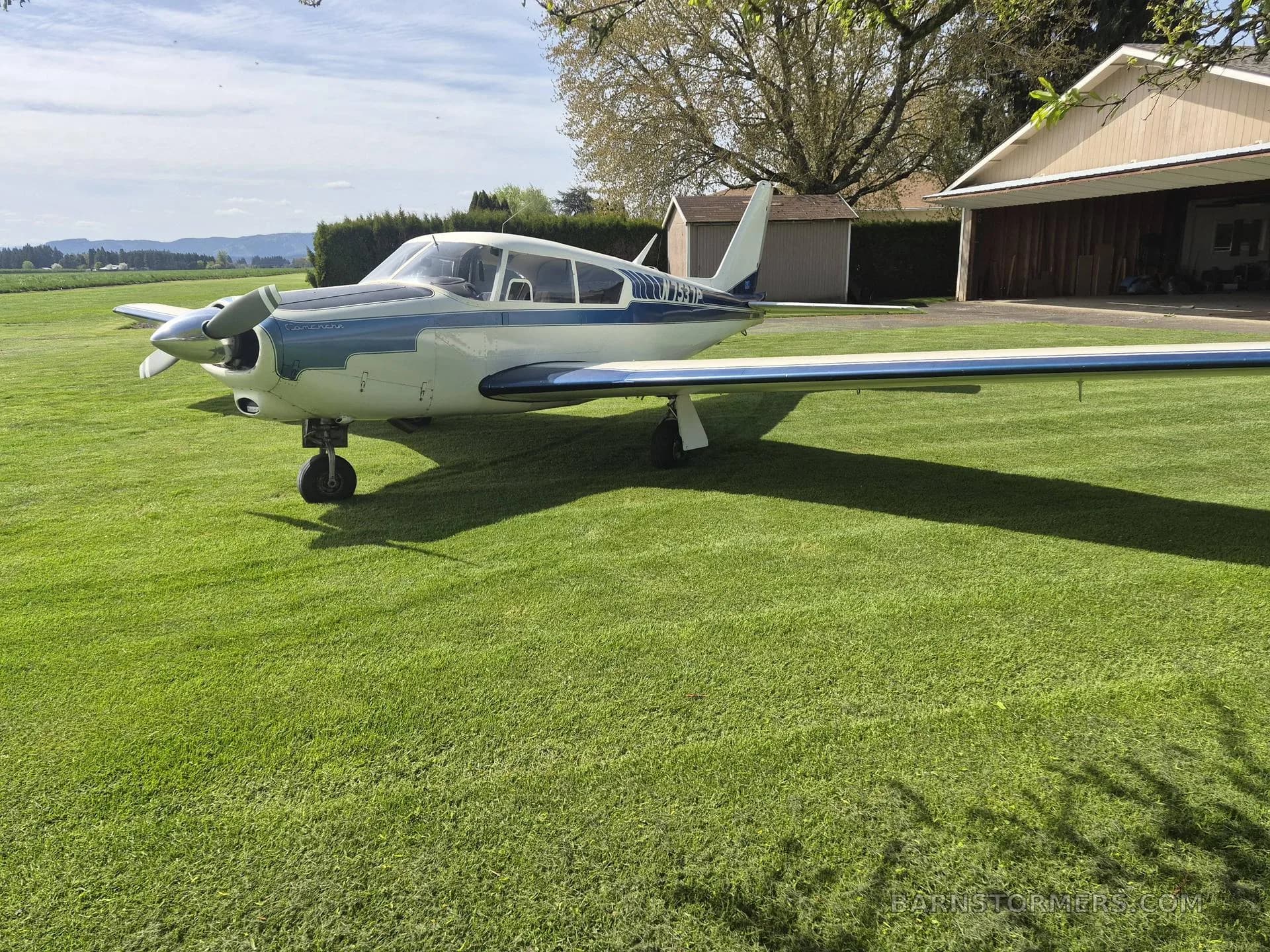 A white and blue single-engine propeller aircraft parked on a green grassy field with a hangar in the background.