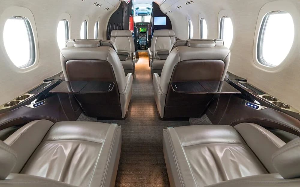 The interior cabin of a private jet, looking forward towards the cockpit, showing light brown leather seats and deployed tables.