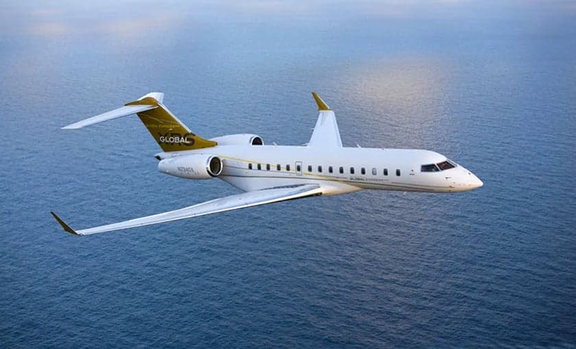 A white and gold Bombardier Global Express business jet flies over a calm blue ocean.